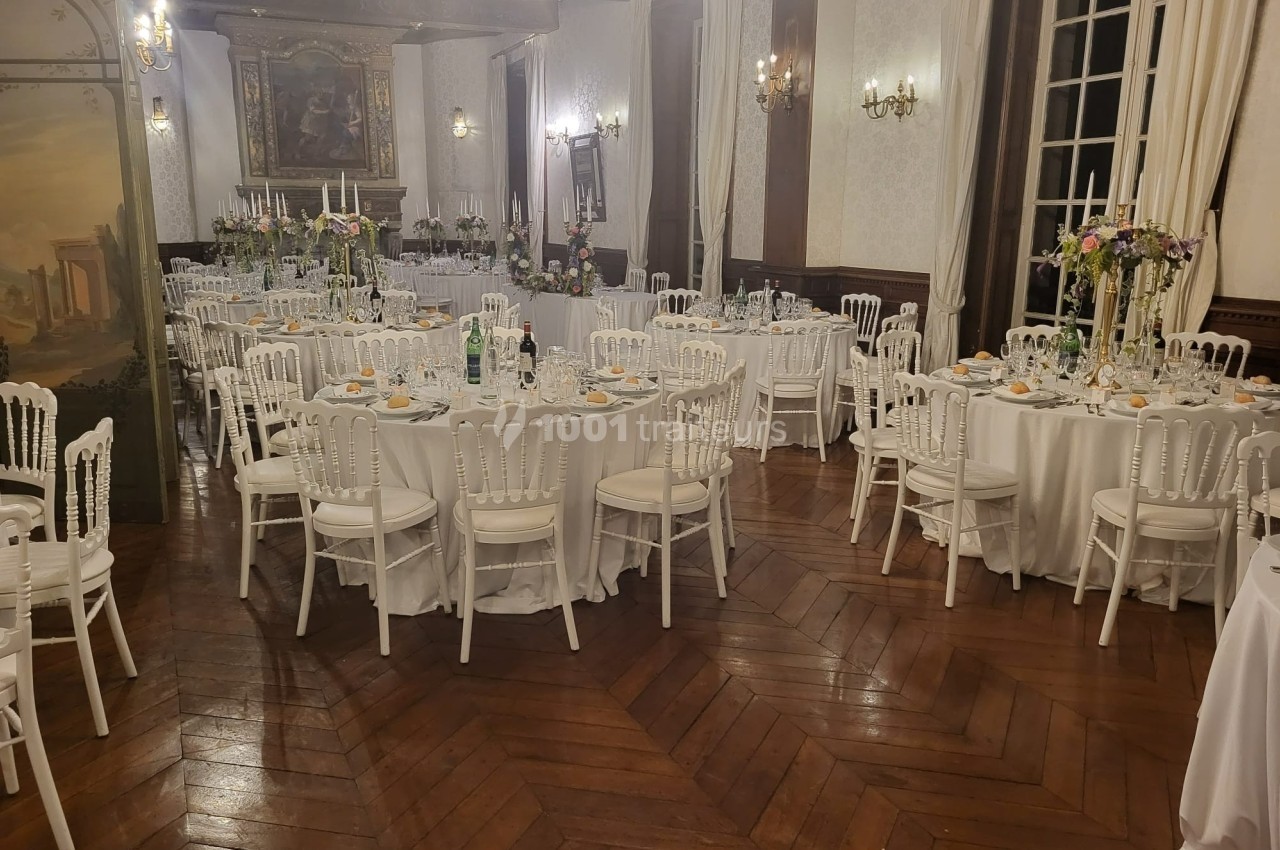 Salle de réception élégante avec tables rondes dressées, chaises blanches et décorations florales.