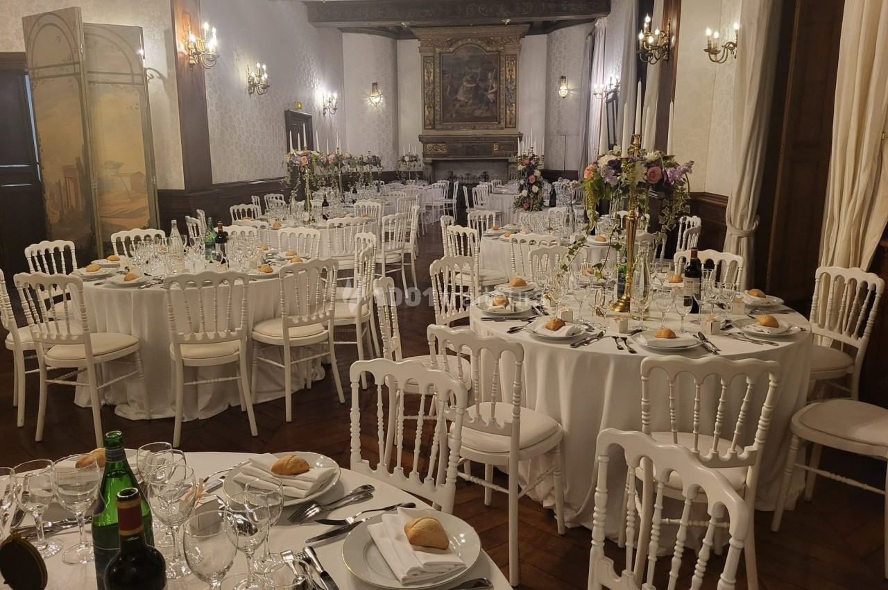 Salle de réception élégante avec tables rondes dressées, décor floral et chaises blanches en bois.