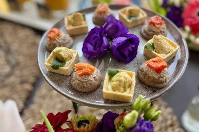 Plateau de bouchées apéritives variées décoré de fleurs violettes, entouré de bouquets colorés et de boissons en arrière…