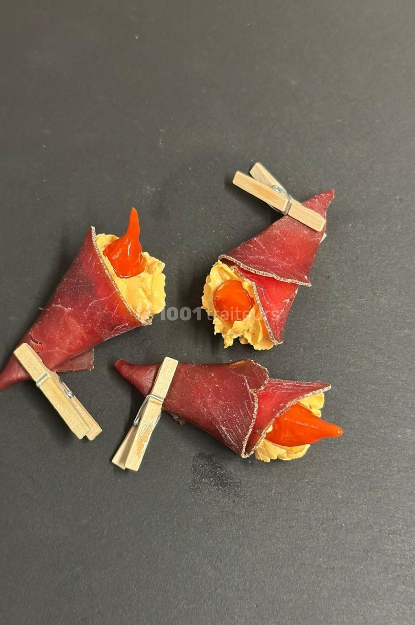 Trois amuse-bouches enroulés dans de la viande séchée, garnis de fromage et décorés de piments rouges, fixés par des pinces.