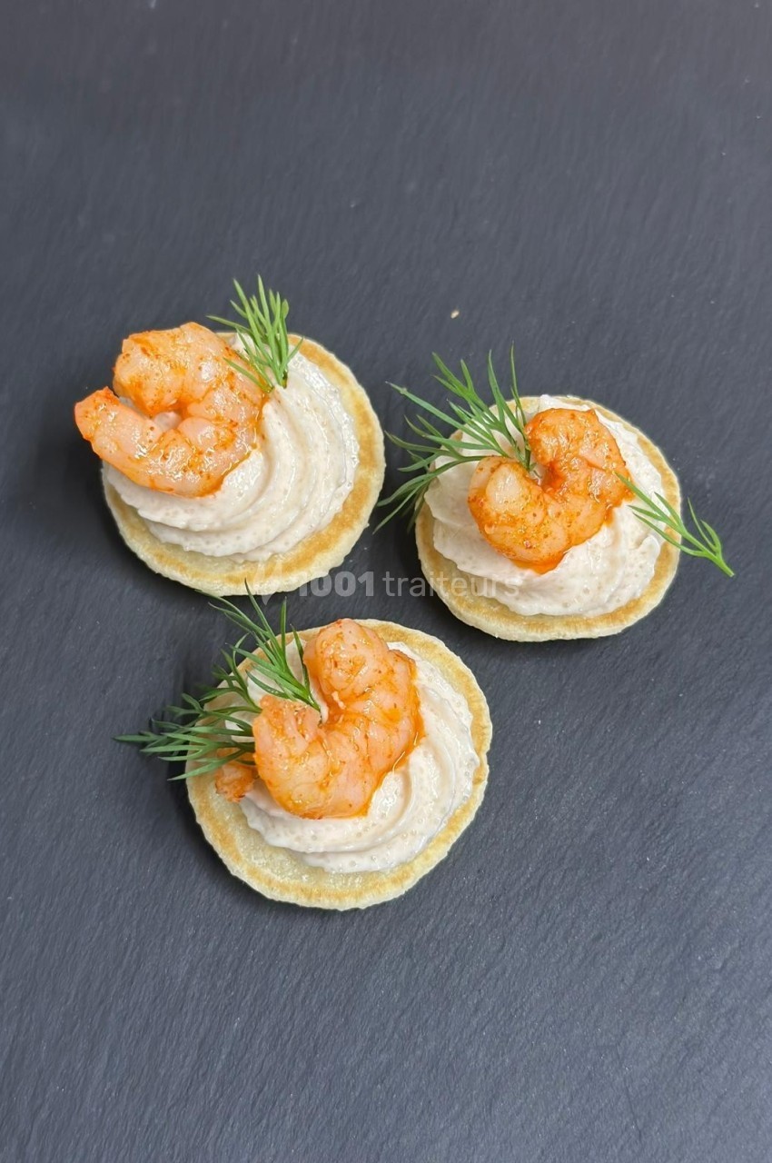 Canapés sur crackers garnis de crème, crevettes épicées et brins d'aneth, présentés sur une ardoise noire.