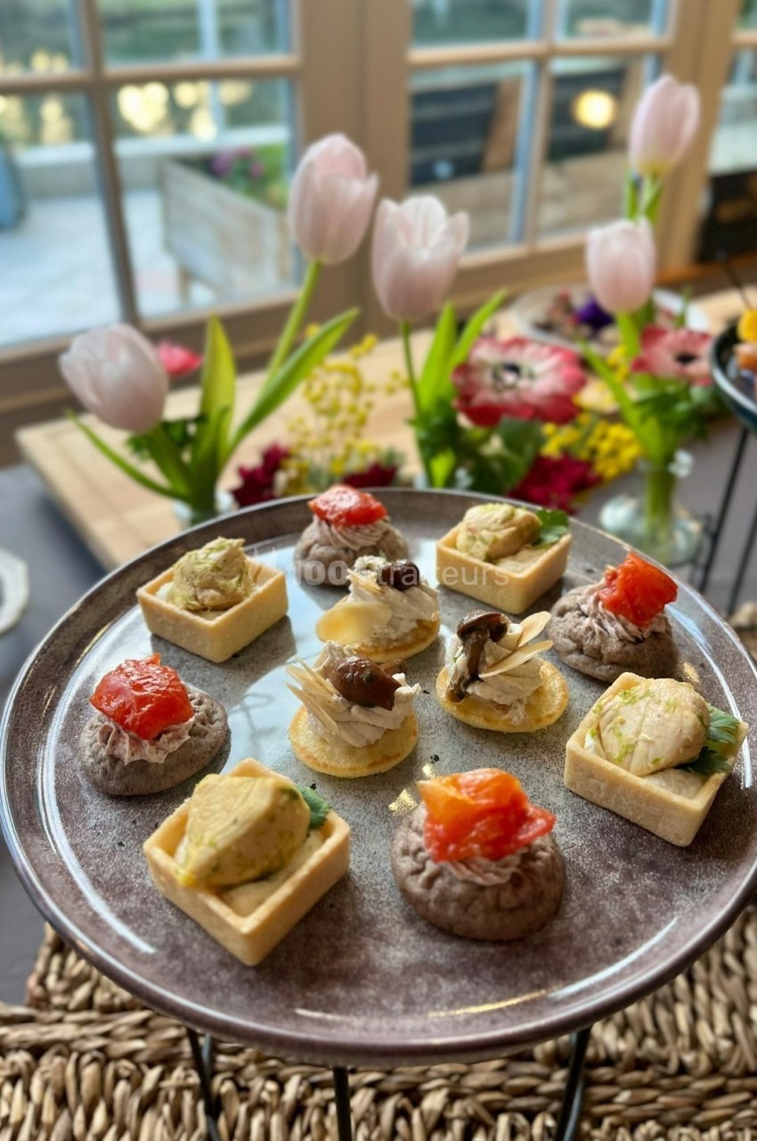 Plateau de bouchées apéritives variées présenté sur une table décorée de fleurs près d'une fenêtre.