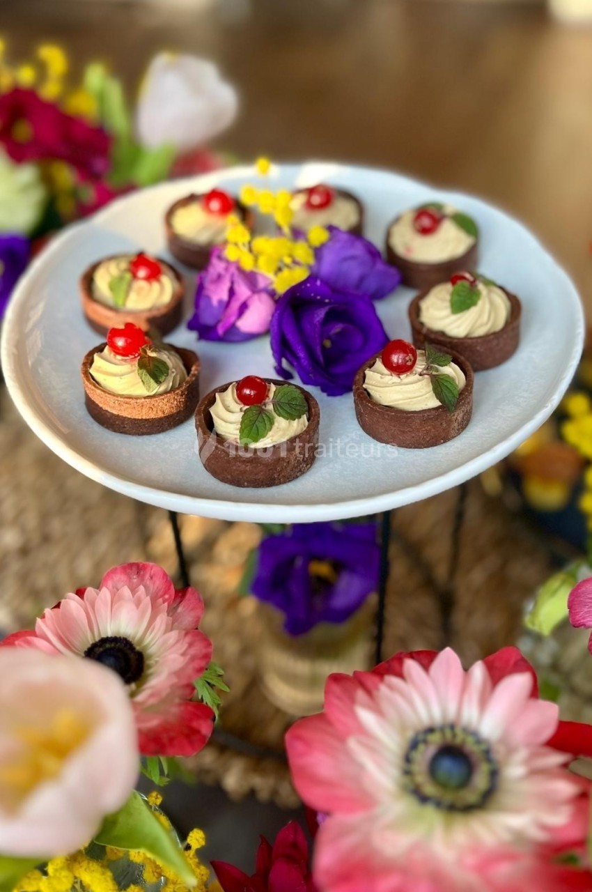 Assortiment de petits desserts chocolatés garnis de crème et cerises, présentés sur un plat entouré de fleurs colorées.
