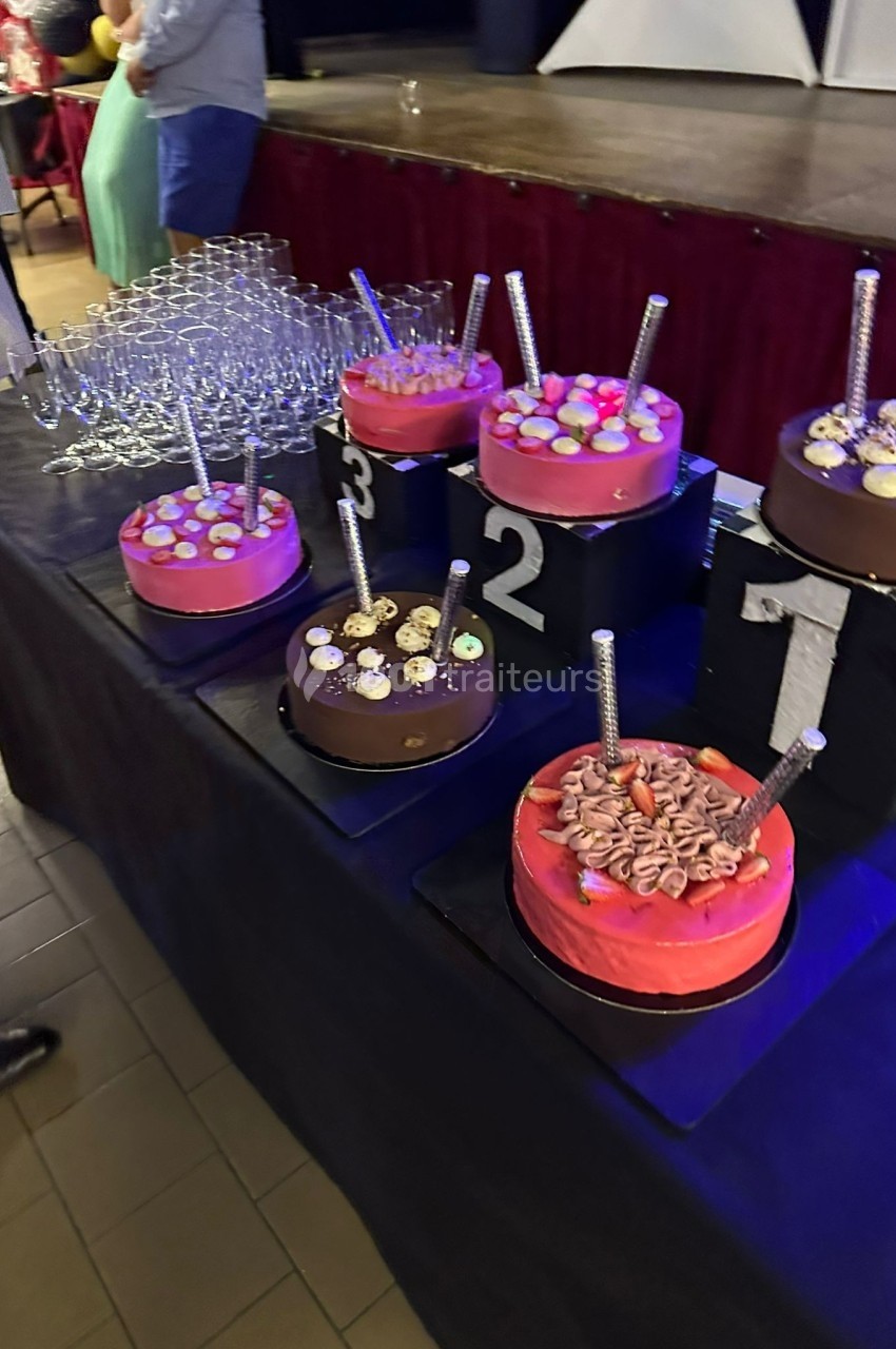 Gâteaux décorés numérotés de 1 à 3 sur une table noire, entourés de verres empilés et d'invités en arrière-plan.