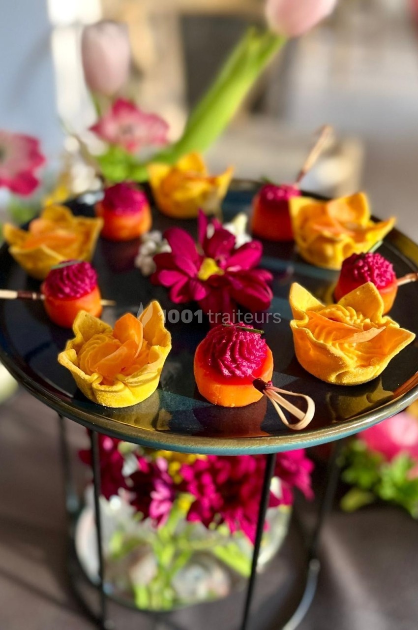 Plateau de bouchées colorées décorées de fleurs comestibles, entouré de fleurs fraîches sur une table.