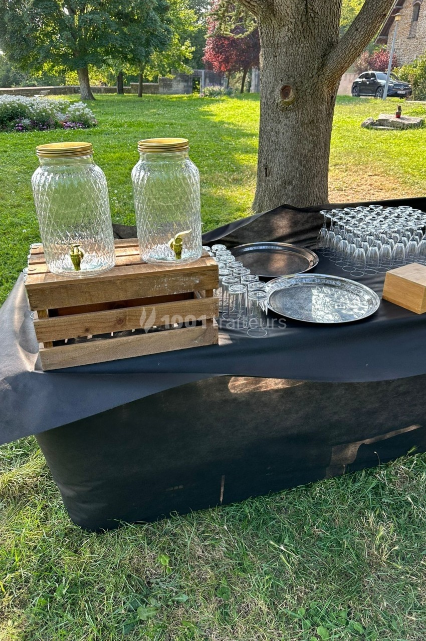 Deux distributeurs en verre sur une table en extérieur, entourés de verres et accessoires de service.