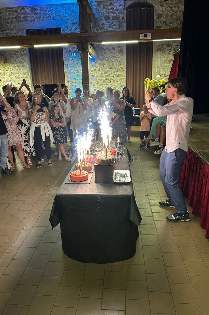 Un homme face à des gâteaux d'anniversaire avec des bougies scintillantes, entouré d'invités dans une salle festive.