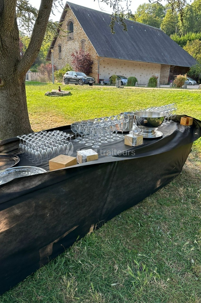 Table extérieure dressée avec des verres, plateaux et seaux à glace, située dans un jardin près d'une grange.