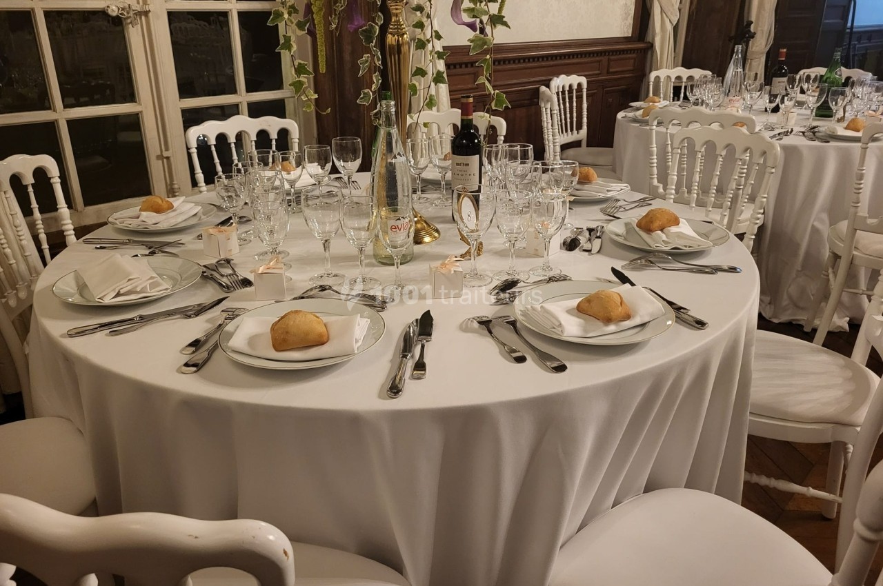 Table ronde dressée pour un repas, avec nappes blanches, couverts, verres, assiettes et décorations florales.