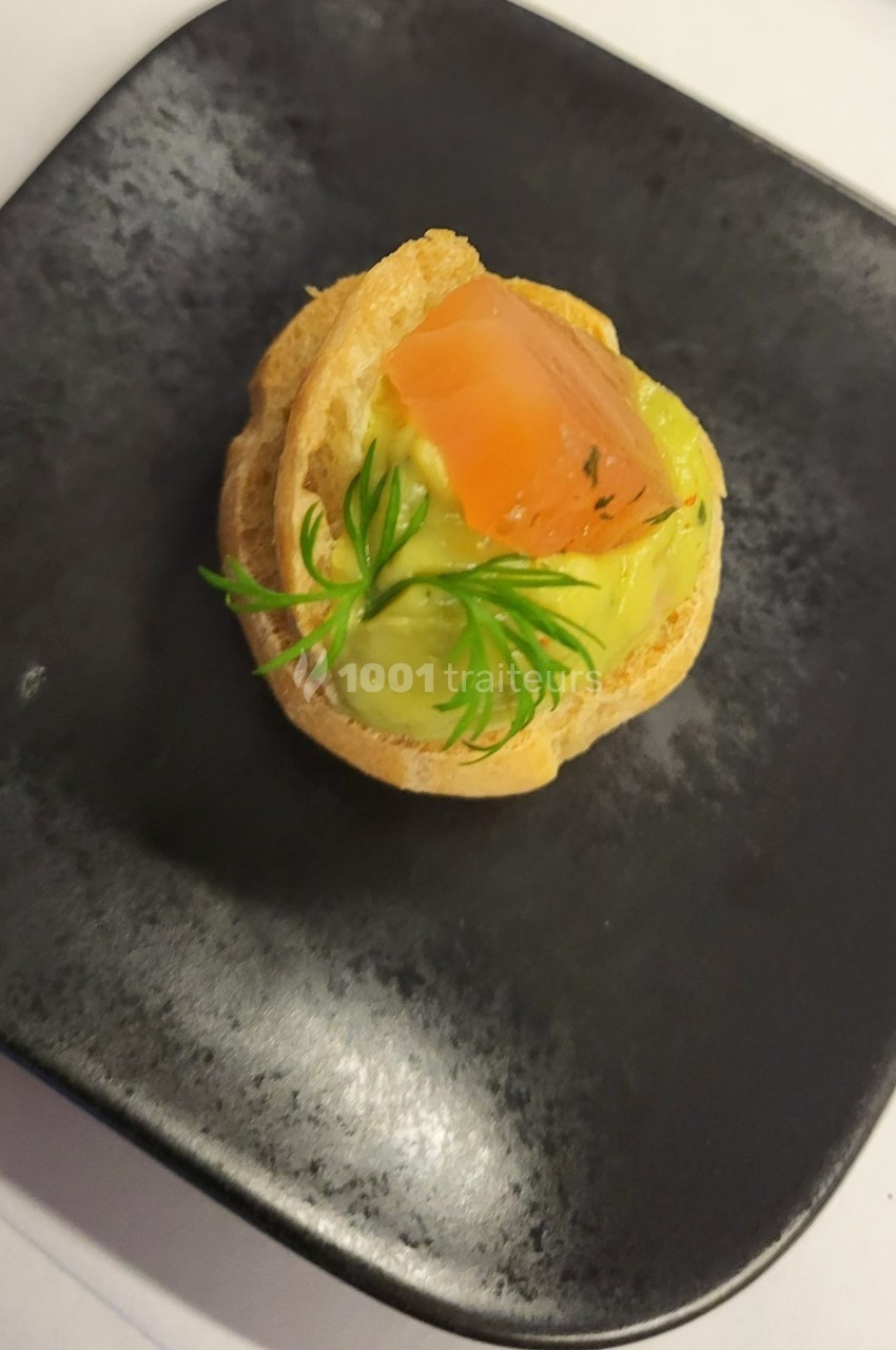 Chou garni de crème à l'avocat, surmonté d'un morceau de saumon fumé et décoré d'aneth, présenté sur une assiette noire.