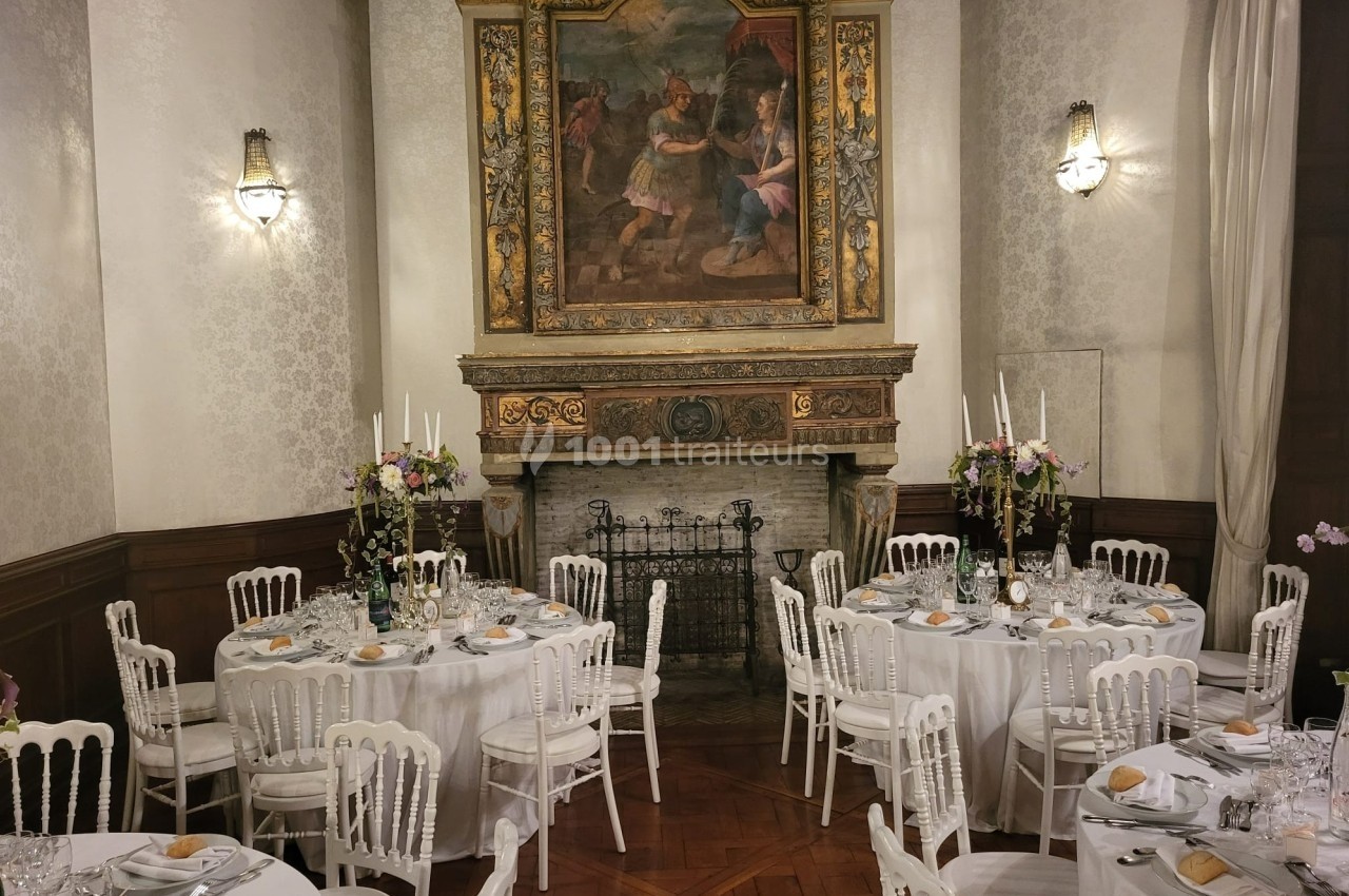 Salle élégante avec tables rondes dressées pour un repas, cheminée ornée et tableau ancien en arrière-plan.