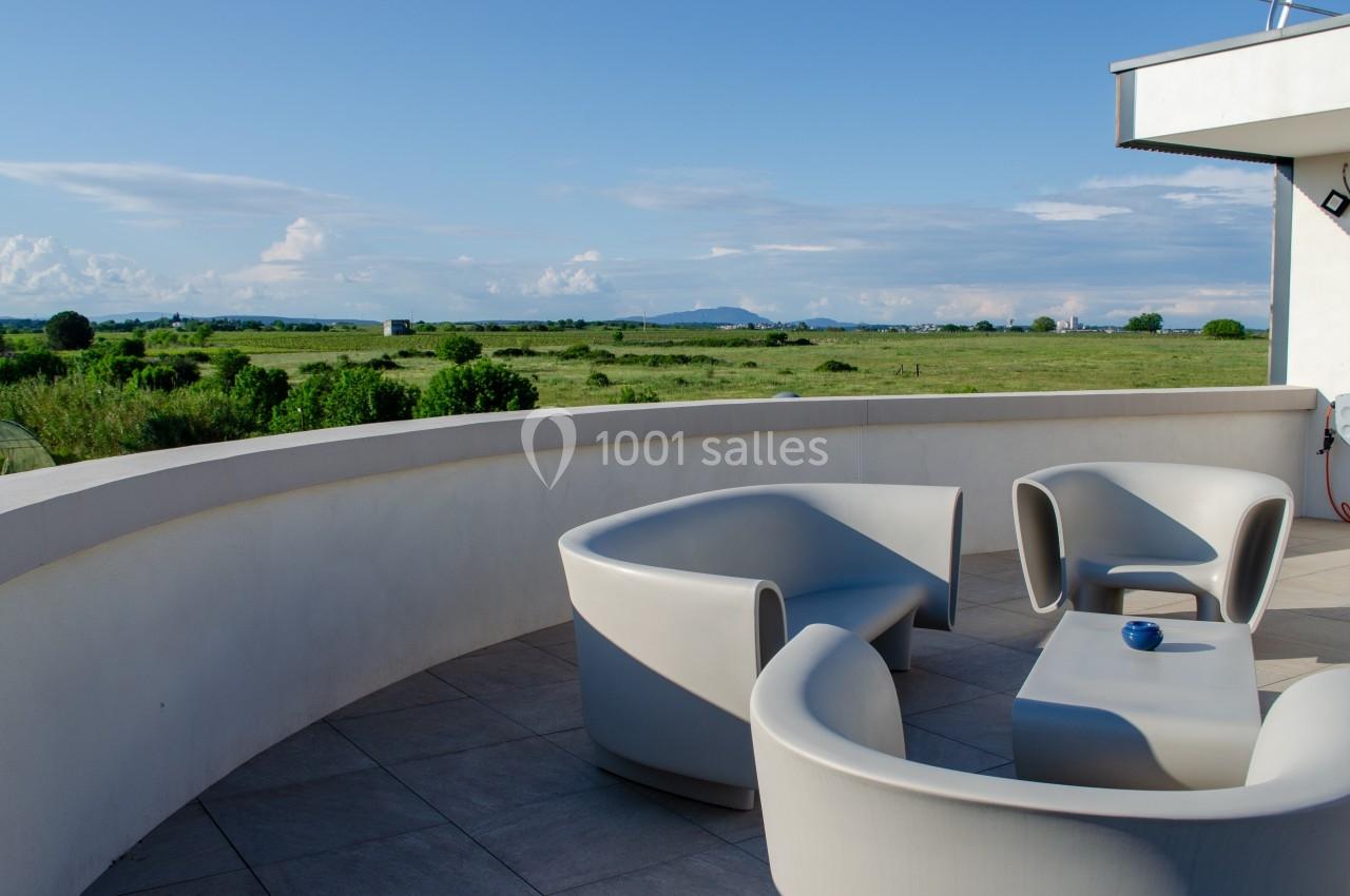 Terrasse avec mobilier moderne en résine grise, offrant une vue dégagée sur une plaine verdoyante et un ciel bleu.
