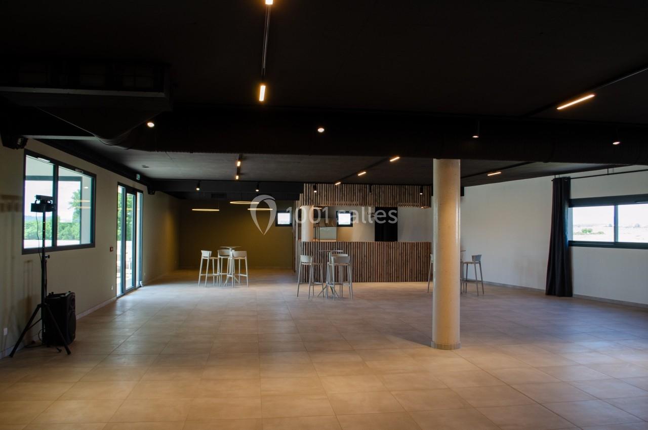 Salle vide avec sol en carrelage, murs clairs, plafond sombre, grandes fenêtres et quelques tables hautes avec tabourets.