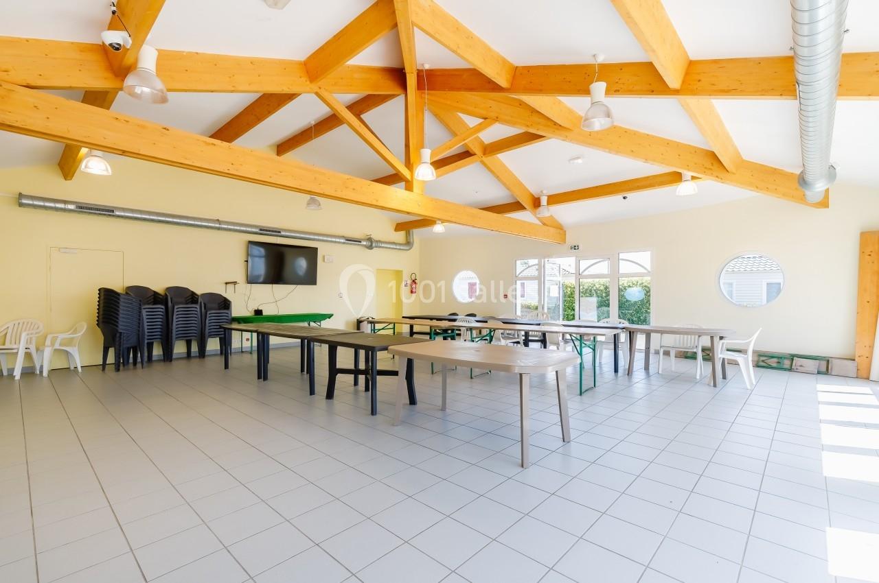 Salle lumineuse avec poutres apparentes, tables et chaises empilées, carrelage clair et fenêtres donnant sur l'extérieur.