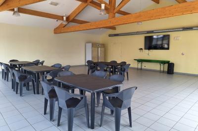 Salle lumineuse avec tables et chaises en plastique noir, plafond en poutres apparentes et écran mural.