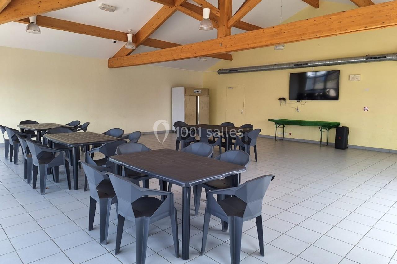 Salle lumineuse avec tables et chaises en plastique noir, plafond en poutres apparentes et écran mural.