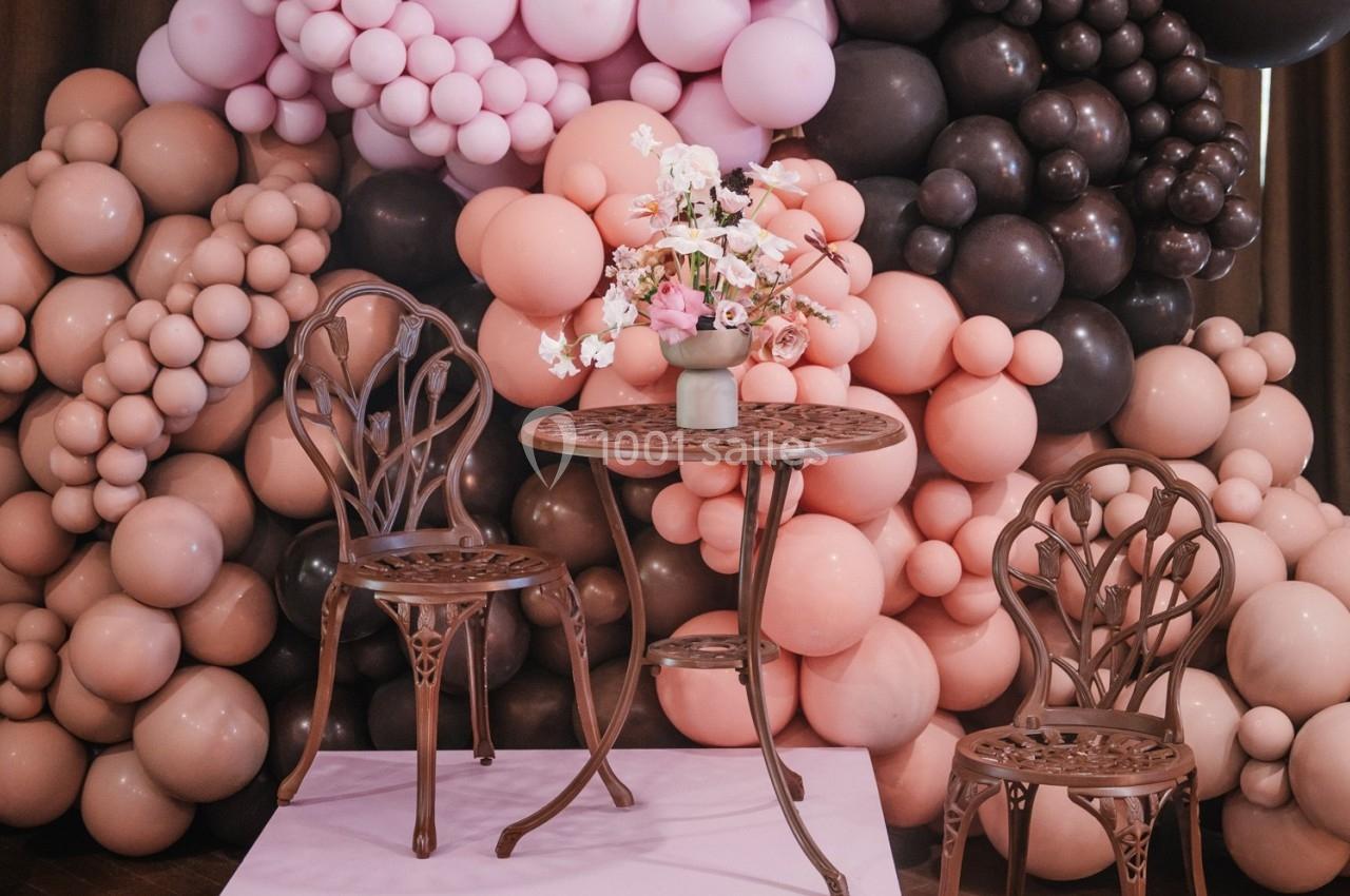 Deux chaises et une table en métal décoratif devant un mur de ballons roses, bruns et beiges, avec un vase de fleurs.