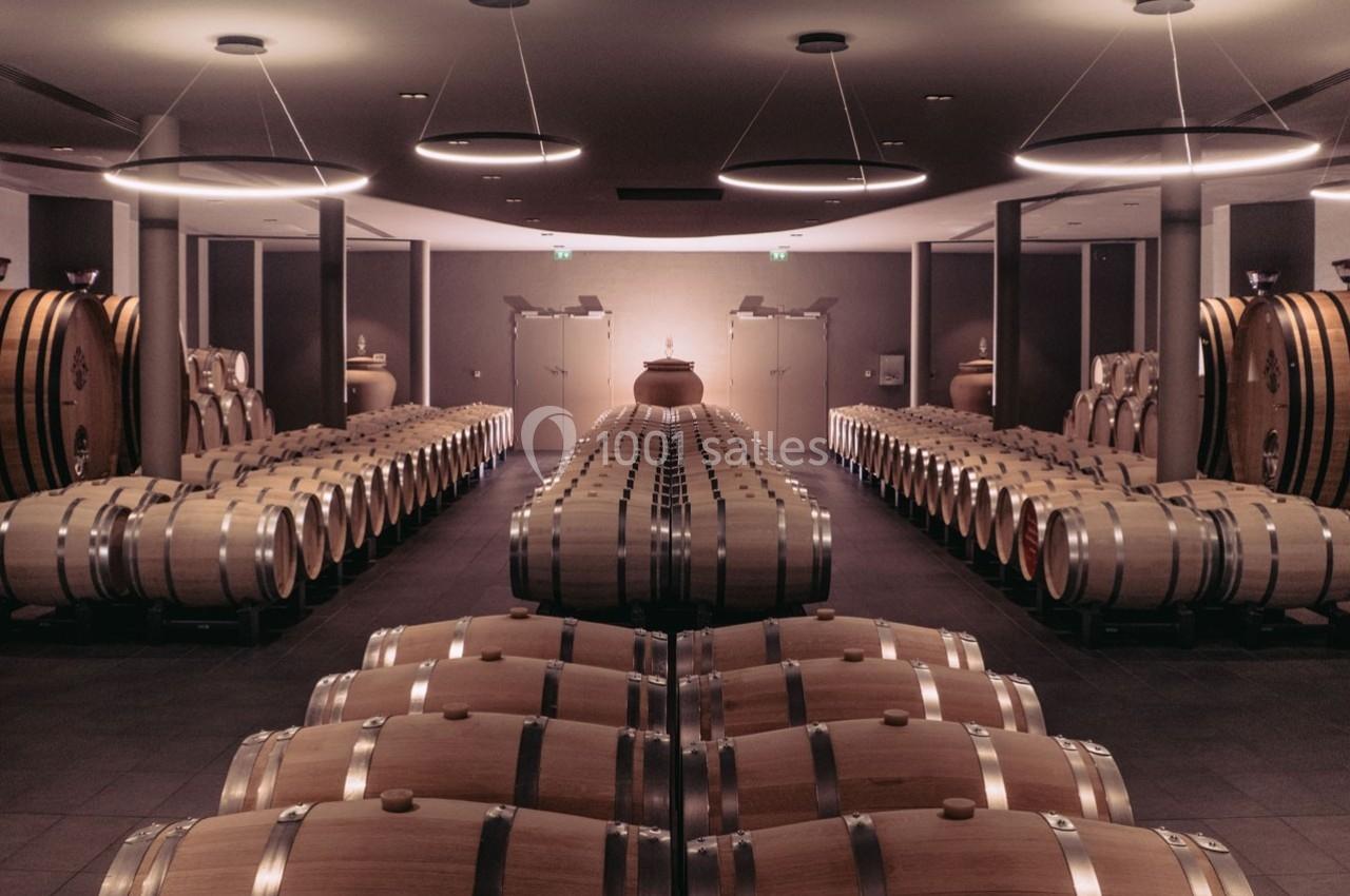 Rangées de fûts en bois alignés dans une cave à vin éclairée par des lumières suspendues.