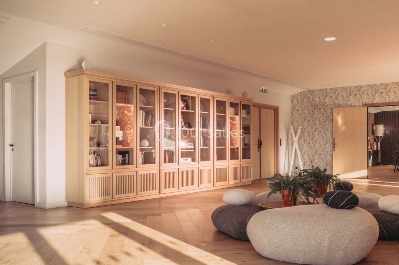 Salle lumineuse avec parquet en bois, bibliothèque vitrée, poufs ronds et plantes décoratives.