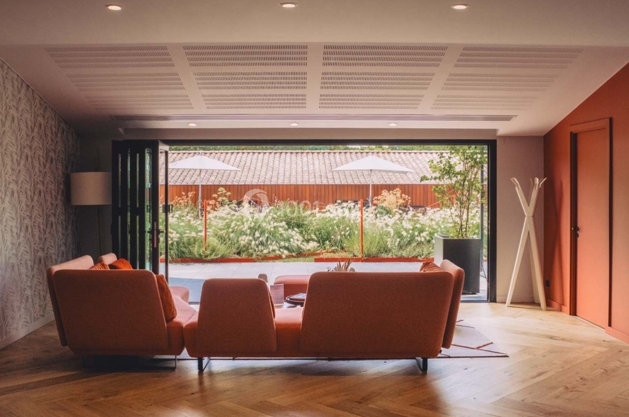 Salon moderne avec canapé orange, parquet en bois, grandes baies vitrées ouvertes sur un jardin fleuri et terrasse.