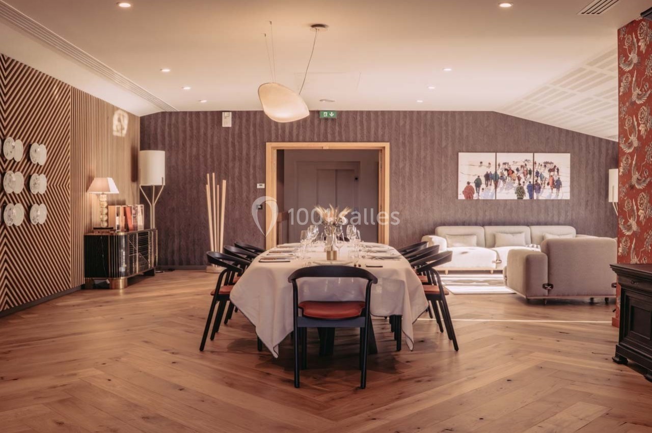 Salle à manger élégante avec une grande table dressée, entourée de chaises noires, et un coin salon en arrière-plan.