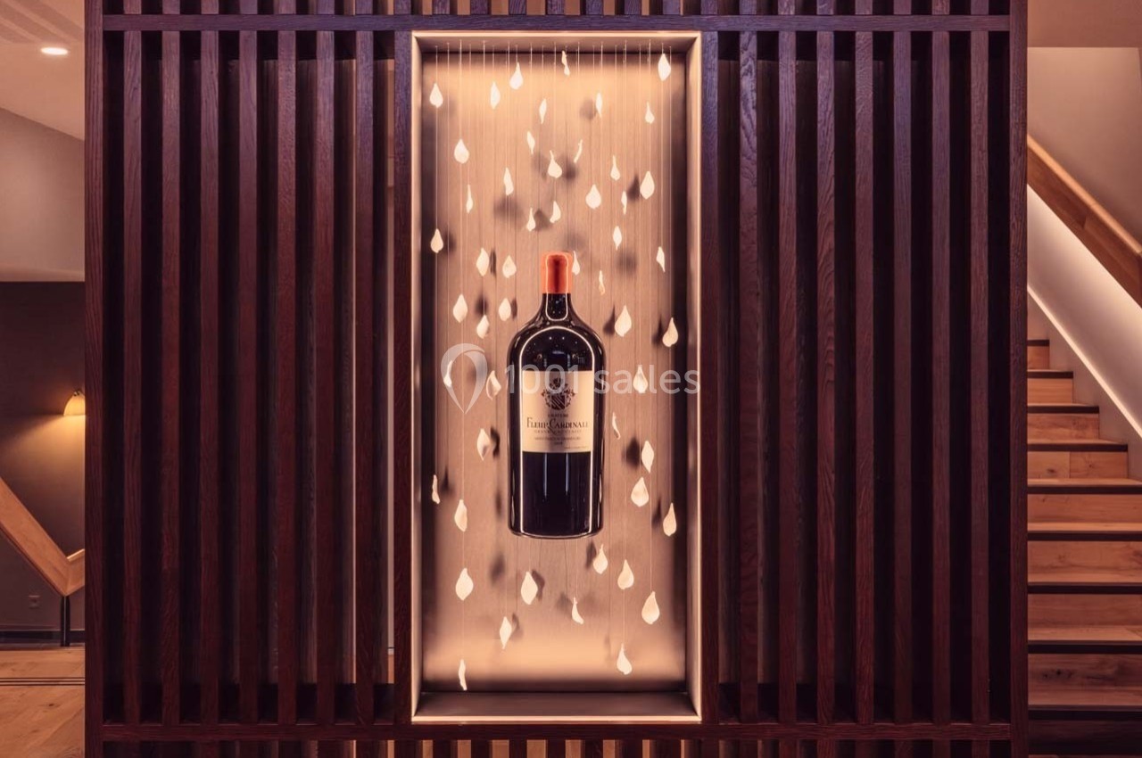 Bouteille de vin rouge exposée dans une niche éclairée, entourée de gouttes suspendues, encadrée par des lattes en bois.