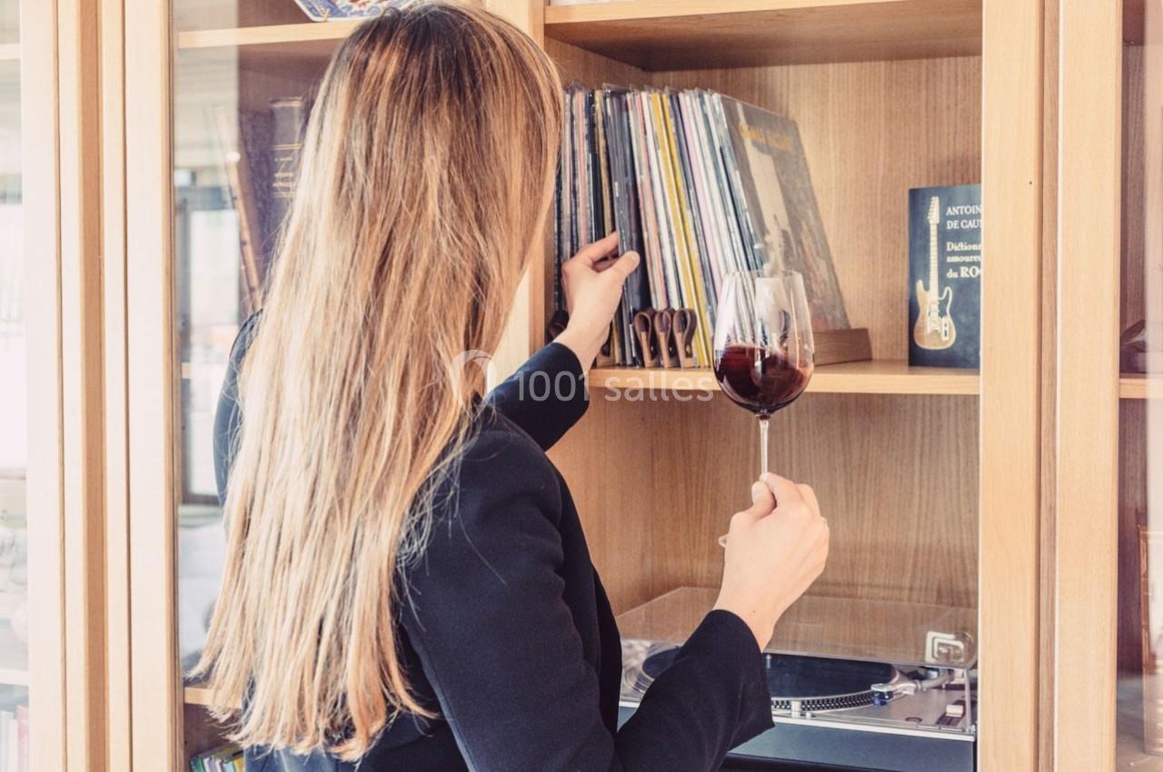 Une femme de dos, tenant un verre de vin rouge, choisit un disque sur une étagère en bois.