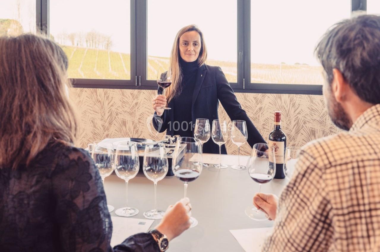 Une femme présente une dégustation de vin à deux personnes assises, avec des verres et une bouteille sur la table.