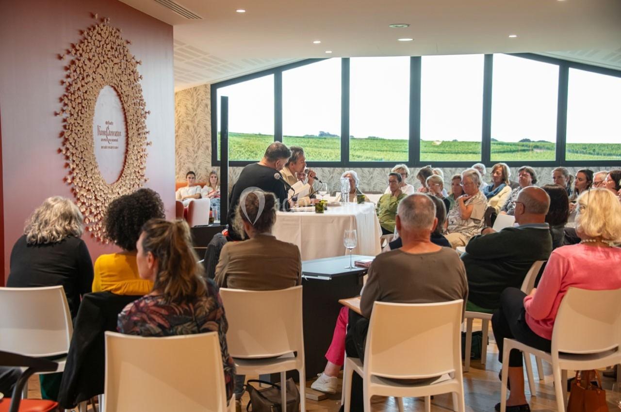Un groupe de personnes assises assiste à une conférence dans une salle lumineuse avec vue sur des vignes.