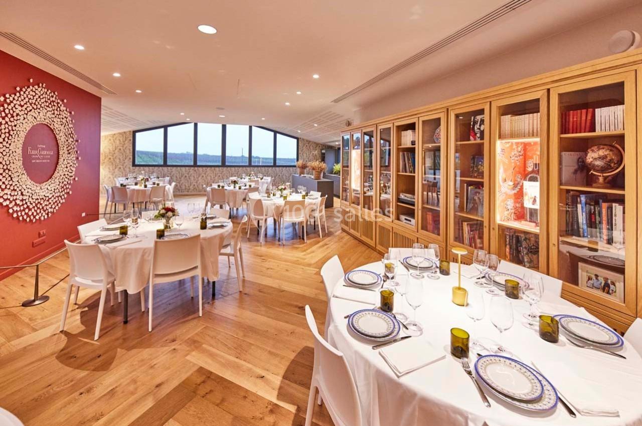 Salle de réception lumineuse avec tables dressées, parquet en bois, mur rouge décoratif et étagères remplies de livres.