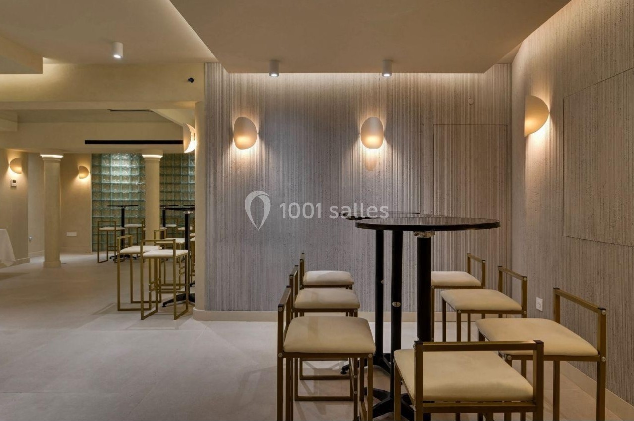 Salle moderne avec des murs texturés, des appliques lumineuses, des tables hautes noires et des chaises à structure dorée.