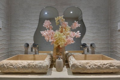 Deux lavabos en pierre avec un vase de fleurs roses et jaunes, placés devant un miroir ondulé.