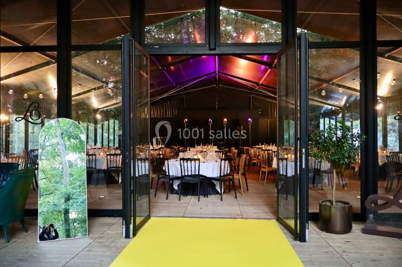 Salle de réception vitrée avec tables dressées, vue depuis l'entrée ouverte sur un tapis jaune.