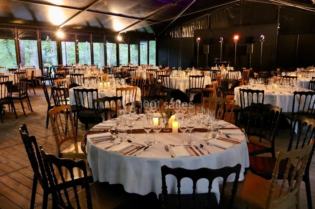 Salle de réception avec tables rondes dressées, nappes blanches, bougies et chaises en bois dans un espace couvert.