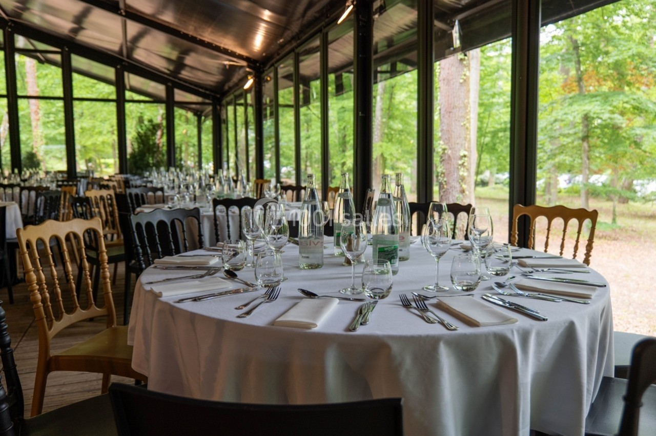 Salle de réception avec tables rondes dressées, nappes blanches, couverts et vue sur un jardin verdoyant à travers de…