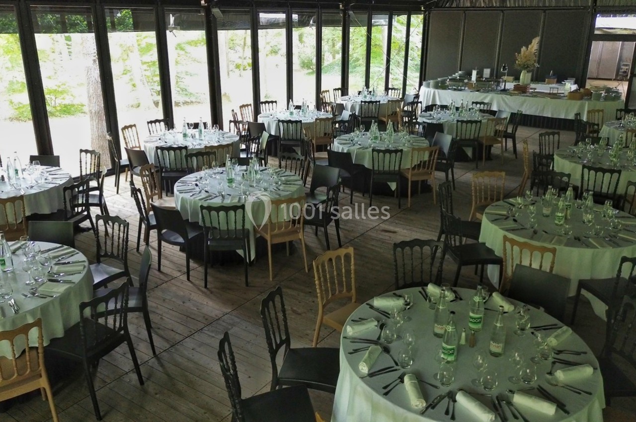 Salle de réception lumineuse avec tables rondes dressées, entourées de chaises, et grandes baies vitrées donnant sur un…