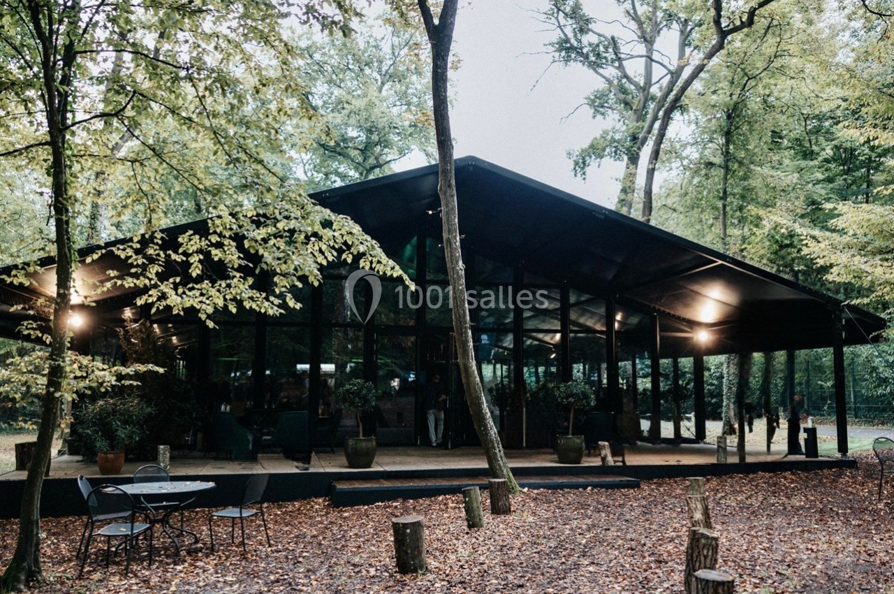 Pavillon moderne en verre et métal entouré d'arbres dans une forêt, avec des tables extérieures sur un sol couvert de…