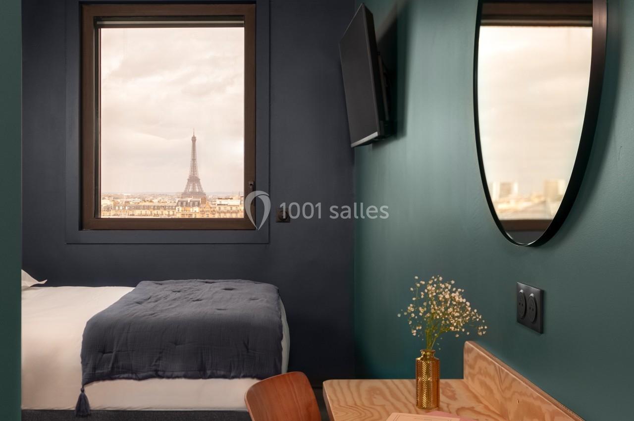 Chambre avec bureau en bois, miroir rond, lit et vue sur la Tour Eiffel par une fenêtre.