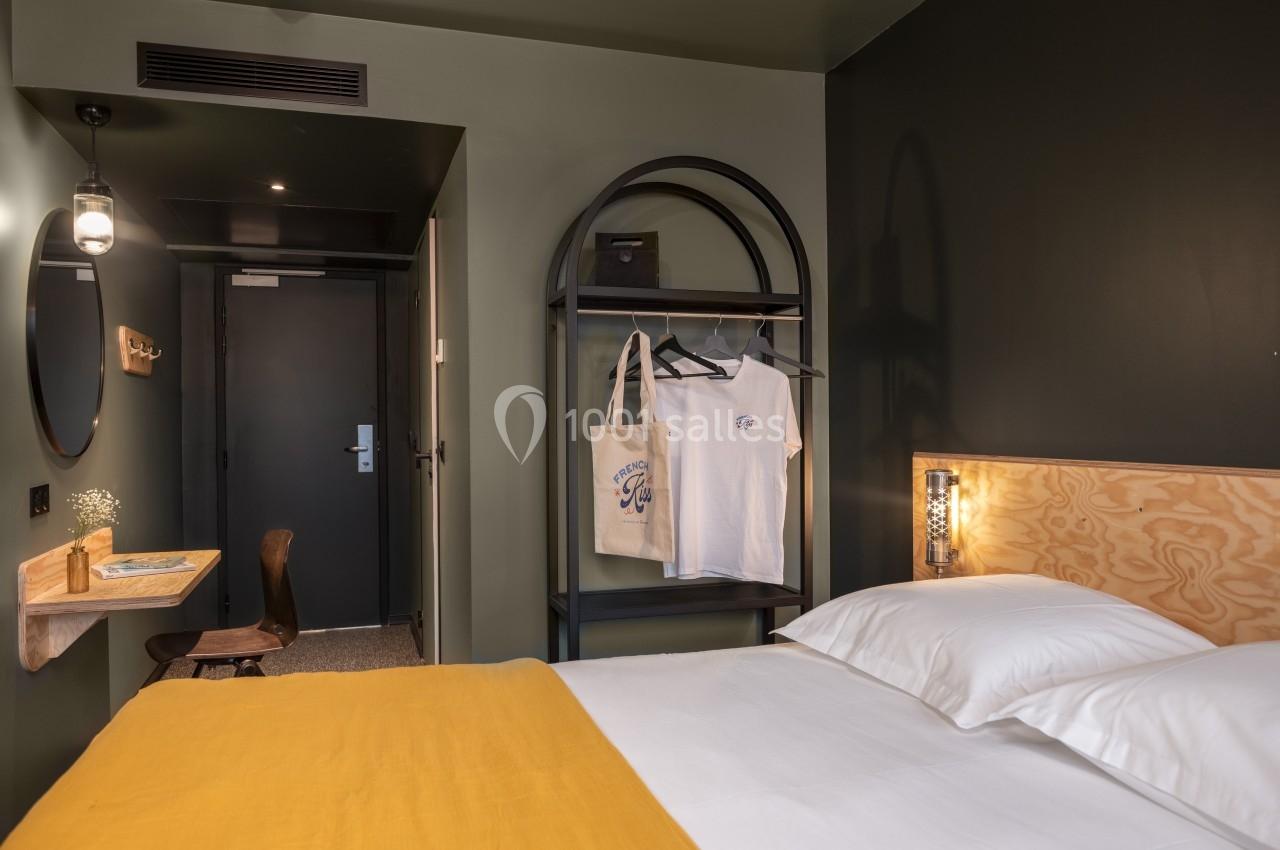 Chambre d'hôtel avec lit double, mur vert, bureau en bois, portant avec vêtements et porte d'entrée au fond.