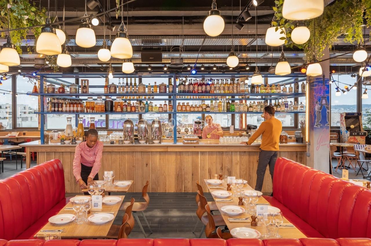 Salle de restaurant moderne avec banquettes rouges, tables dressées, bar en arrière-plan et personnel en préparation.