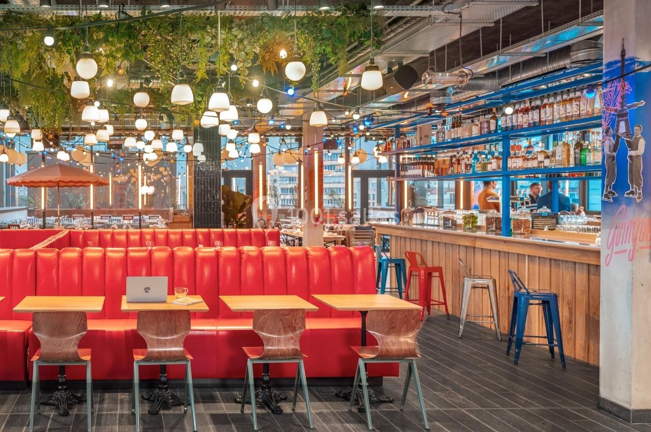 Salle de restaurant moderne avec banquettes rouges, tables en bois, bar éclairé et décoration végétale suspendue.