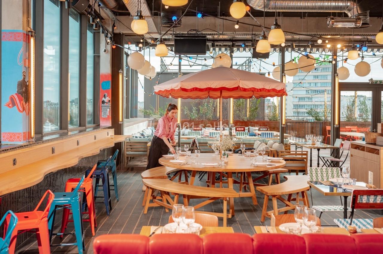 Salle de restaurant lumineuse avec grandes baies vitrées, mobilier coloré et parasol central décoratif.