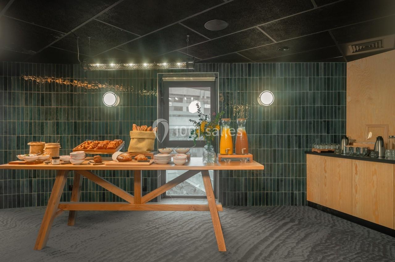 Table en bois dressée avec viennoiseries, pains, jus d'orange et vaisselle, dans une salle aux murs carrelés verts.