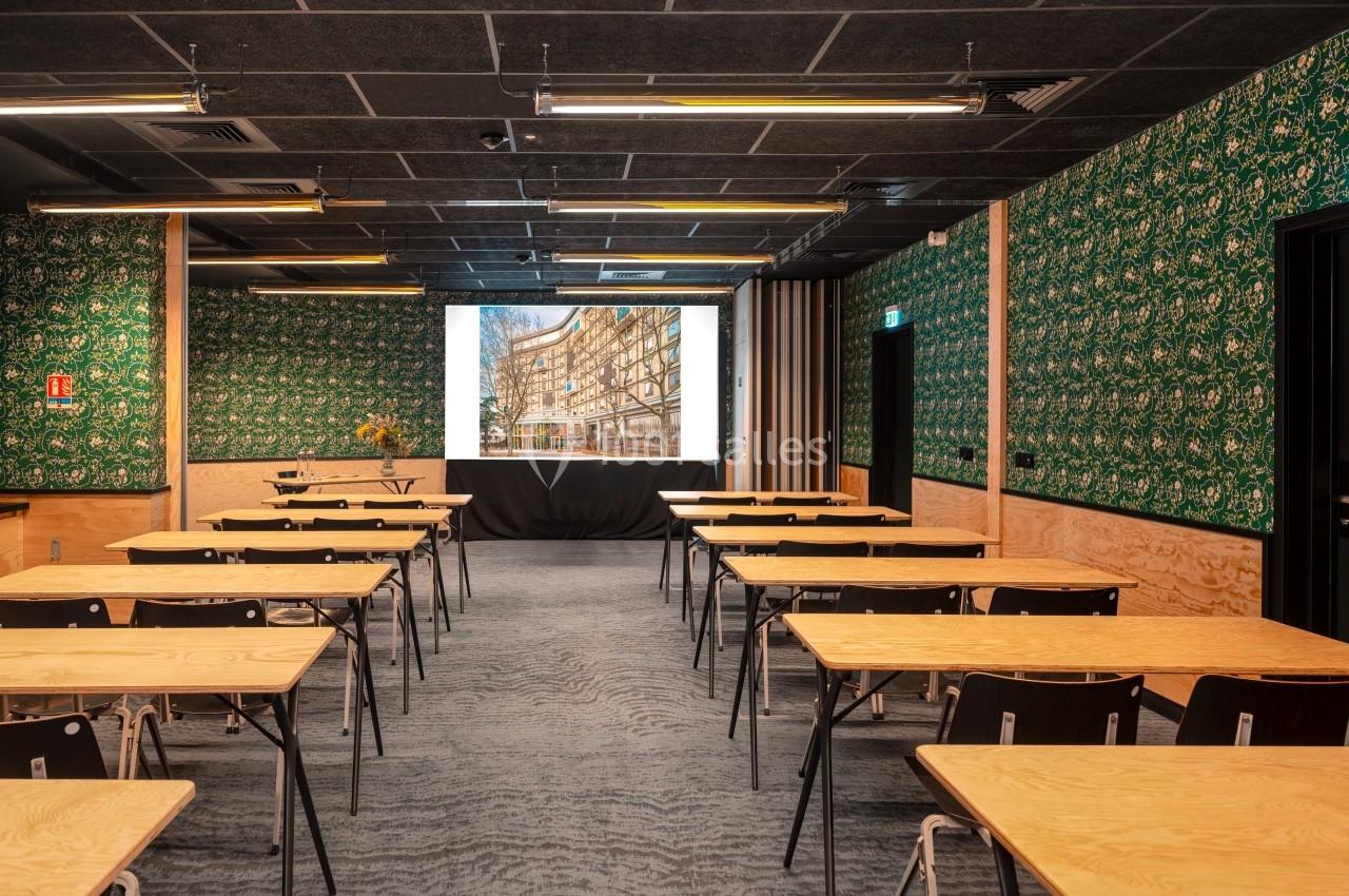 Salle de conférence avec tables en bois clair, chaises noires et écran de projection allumé, décor mural vert à motifs.
