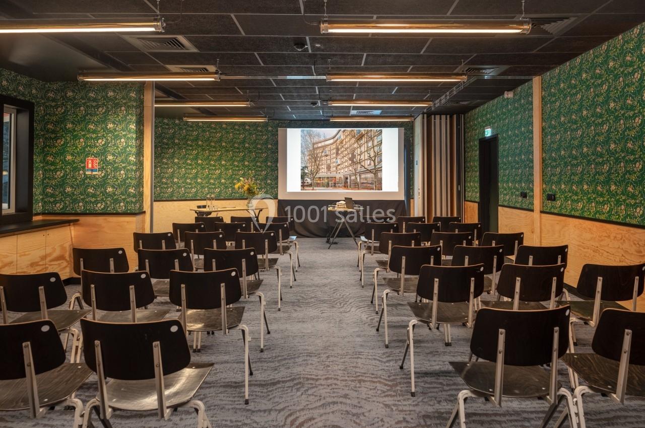 Salle de conférence avec des rangées de chaises noires, un écran de projection allumé et des murs décorés de motifs verts.