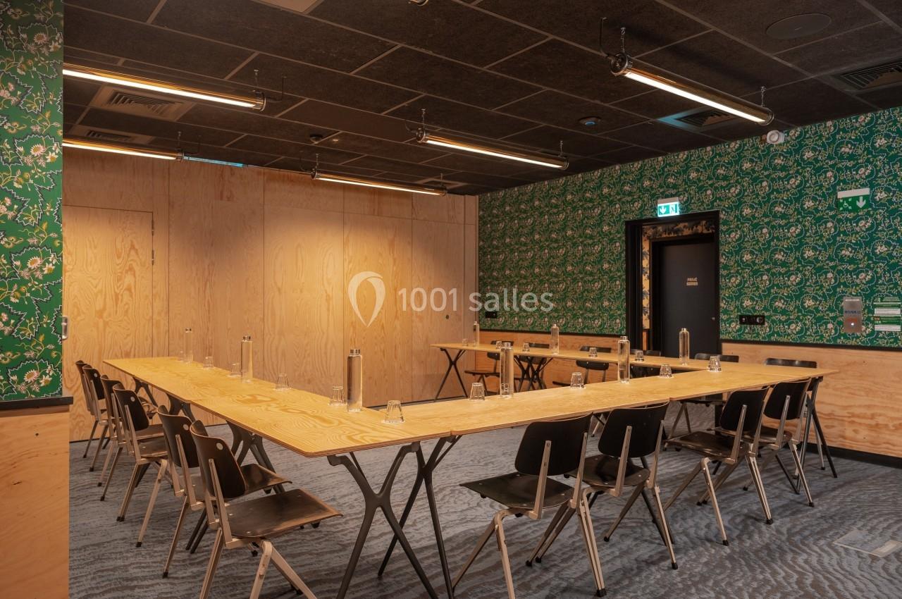 Salle de réunion avec tables en bois disposées en U, chaises noires et murs décorés de bois et de papier peint vert.