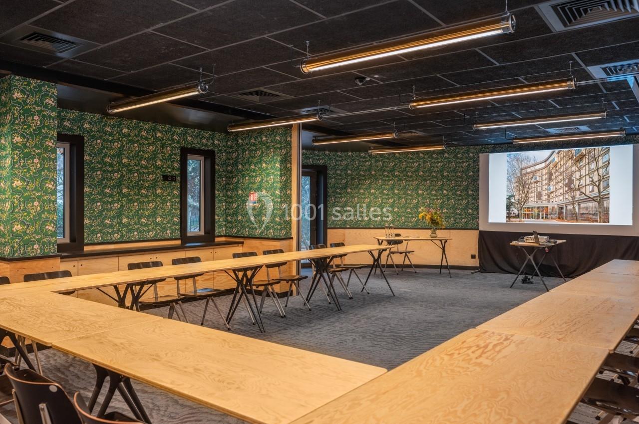 Salle de réunion avec tables en bois disposées en U, écran de projection allumé et murs décorés de papier peint vert.