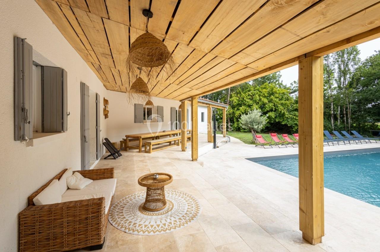 Terrasse couverte avec mobilier en bois et rotin, vue sur une piscine entourée de chaises longues colorées et végétation.