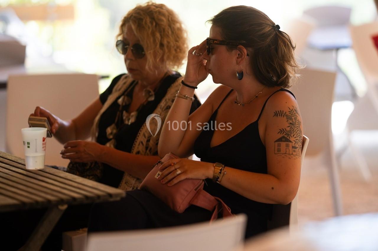 Deux femmes assises à une table en extérieur, discutant, l'une tenant un objet et l'autre ajustant ses lunettes.