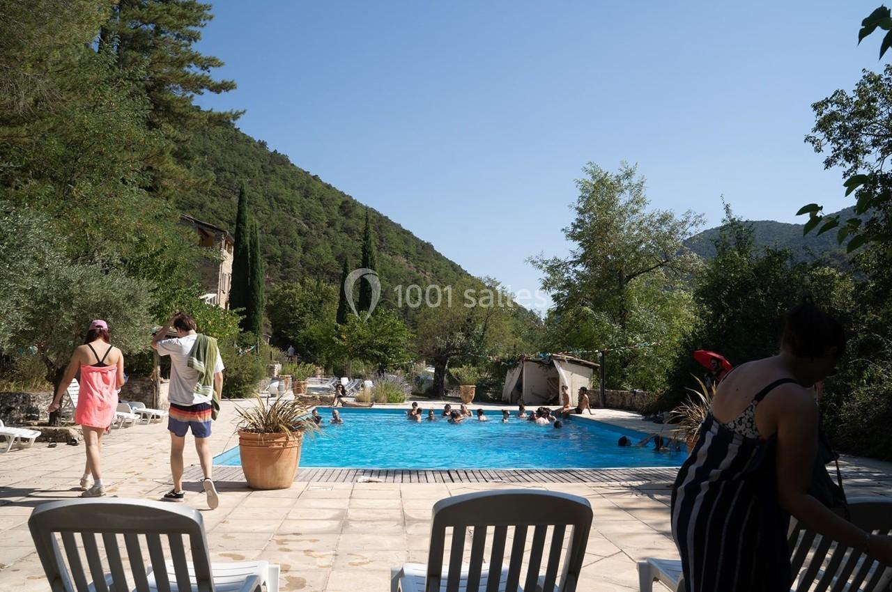 Piscine extérieure entourée de chaises longues, avec des baigneurs et un paysage de collines verdoyantes en arrière-plan.